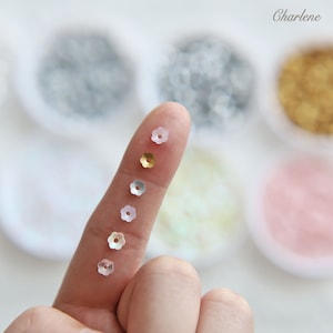 May include: A variety of flower-shaped sequins in different sizes and colors, including gold, silver, and white. The sequins are arranged in a line on a finger.