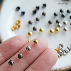 May include: Assortment of small metal studs in gold, silver, and black. The studs have a round head and three prongs. The studs are displayed on a white surface.