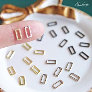 Pode incluir: Conjunto de pequenos anéis metálicos retangulares em ouro, prata e preto. Os anéis estão dispostos sobre uma superfície branca com um fundo dourado.