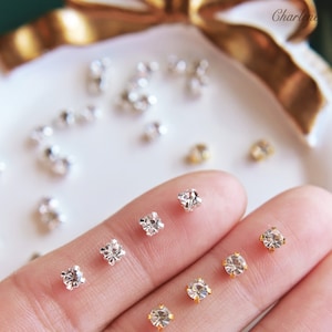 以下が含まれることがあります： さまざまなサイズのクリアラインストーンの詰め合わせ。指と白い皿の上に置かれています。ラインストーンは銀色の裏地が付いています。