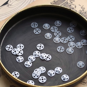 May include: A black tray filled with white plastic snap fasteners. The fasteners are in various sizes and are arranged in a random pattern.