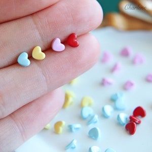 May include: An assortment of small heart-shaped buttons in various colours, including light blue, yellow, pink, and red. The buttons are arranged in a row on a finger, with a larger collection of buttons scattered on a white surface.