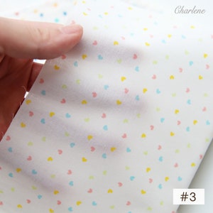 May include: White fabric with a scattered pattern of small pink, blue, and yellow hearts. The fabric is being held in a hand and the number #3 is visible in the bottom right corner.