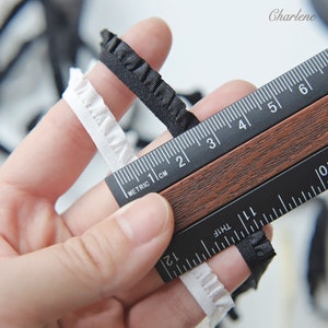 May include: Two pieces of ruffled trim, one white and one black, are measured with a ruler. The ruler is marked in both inches and centimeters. The white trim is 1 inch wide and the black trim is 1/2 inch wide.