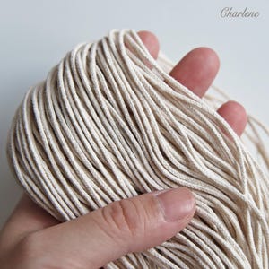 Puede incluir: Una mano sostiene un manojo de cordón de macramé blanquecino. El cordón está bien enrollado y parece estar hecho de una fibra natural y suave. La imagen está bien iluminada, resaltando la textura y el color del cordón. La palabra "Charlene" está en la esquina superior derecha.