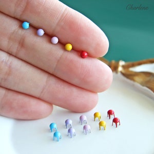 Può includere: Assortimento di puntine metalliche colorate con una varietà di colori tra cui rosso, giallo, blu e viola. Le puntine sono disposte in una linea su una superficie bianca.