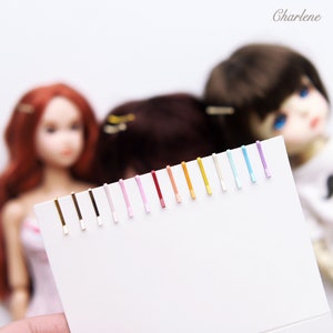 May include: A set of 18 colorful hair clips with gold metal clasps. The clips are arranged in a row on a white surface. The colors include gold, black, white, pink, red, orange, yellow, light blue, dark blue, and purple.