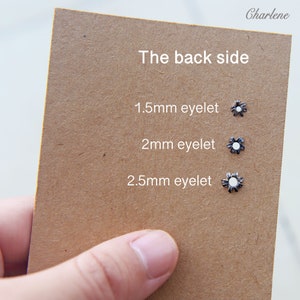 以下が含まれることがあります： 茶色のカードには、1.5mm、2mm、2.5mmとラベル付けされた、白い中心を持つ3つの小さな銀色のハトメが表示されています。「The back side」というテキストが上部にあり、右上に「Charlene」という名前があります。