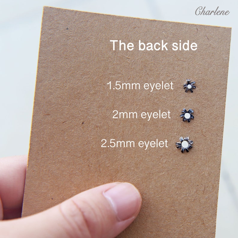 以下が含まれることがあります： 茶色のカードには、1.5mm、2mm、2.5mmとラベル付けされた、白い中心を持つ3つの小さな銀色のハトメが表示されています。「The back side」というテキストが上部にあり、右上に「Charlene」という名前があります。