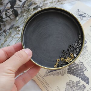 May include: A black metal tray with a gold rim, filled with small gold and black metal flower-shaped beads.