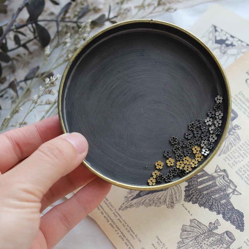 May include: A black metal tray with a gold rim, filled with small gold and black metal flower-shaped beads.