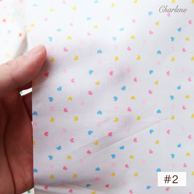 May include: White fabric with a small, scattered pattern of pink, blue and yellow hearts. The fabric is labelled with the number "#2".