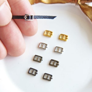 May include: Assortment of eight metal buckles in silver, gold, and black finishes. The buckles are small and rectangular with two holes on each side.