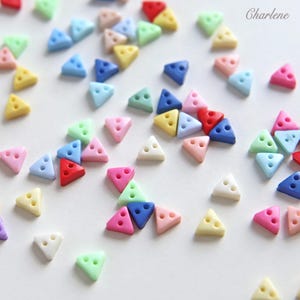 Puede incluir: Una colección de pequeños botones triangulares en varios colores pastel y primarios, cada uno con dos agujeros para coser. Los botones están esparcidos por la superficie y la palabra Charlene está impresa en la esquina superior derecha.