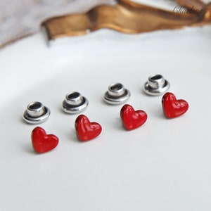 Puede incluir: Cinco botones rojos en forma de corazón con ajustes de metal plateado. Los botones están dispuestos en una fila sobre una superficie blanca.