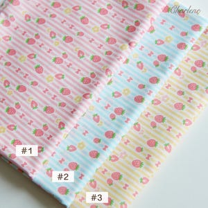 May include: Three fabric swatches with a white background and pink, blue, and yellow stripes. Each swatch has a repeating pattern of small red strawberries, white flowers, and pink bows.