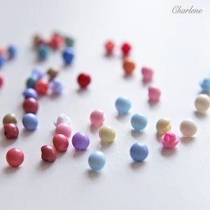 Op de afbeelding: Assortiment van kleurrijke plastic knopen in verschillende tinten roze, blauw, paars en crème. De knopen zijn verspreid over een wit oppervlak.