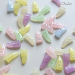 Peut inclure: Une collection de boutons en forme de corne de couleur pastel, dispersés sur une surface blanche. Les boutons sont dans des tons de jaune, rose, vert, violet et bleu. Chaque bouton a deux petits trous. Le mot "Charlene" est dans le coin supérieur droit.