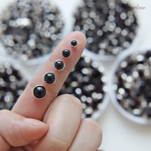 Op de afbeelding: Een verzameling zwarte oorstekers in verschillende maten, gepresenteerd op een vinger. Elke oorbel heeft een glanzende, ronde zwarte steen gezet in een zilverkleurige zetting. De achtergrond toont meer zwarte stenen in kleine witte schaaltjes.