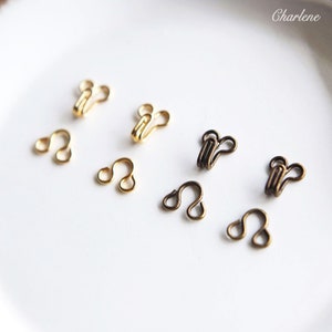 May include: A close-up of eight gold and bronze hook and eye closures. The hooks and eyes are arranged in two rows on a white surface.