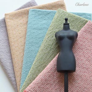 May include: A matte black mannequin torso stands in front of five folded fabric swatches in shades of grey, tan, light blue, sage green, and dusty rose. Each fabric features a subtle, repeating textured cross-stitch pattern. The text Charlene is visible.