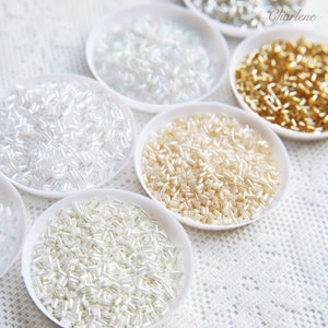 May include: Six small white bowls filled with different colors of seed beads. The beads are all the same shape, but vary in color from white to cream to gold.