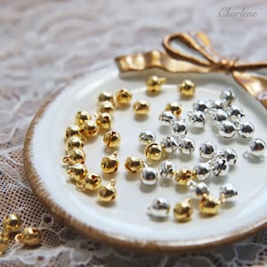 May include: A white plate with a gold rim holds a collection of small, silver and gold jingle bells. The bells are scattered randomly on the plate.