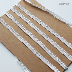 May include: Five white ruffled trim ribbons on a brown background. The ribbons are arranged in parallel lines, with the top ribbon slightly offset from the others.