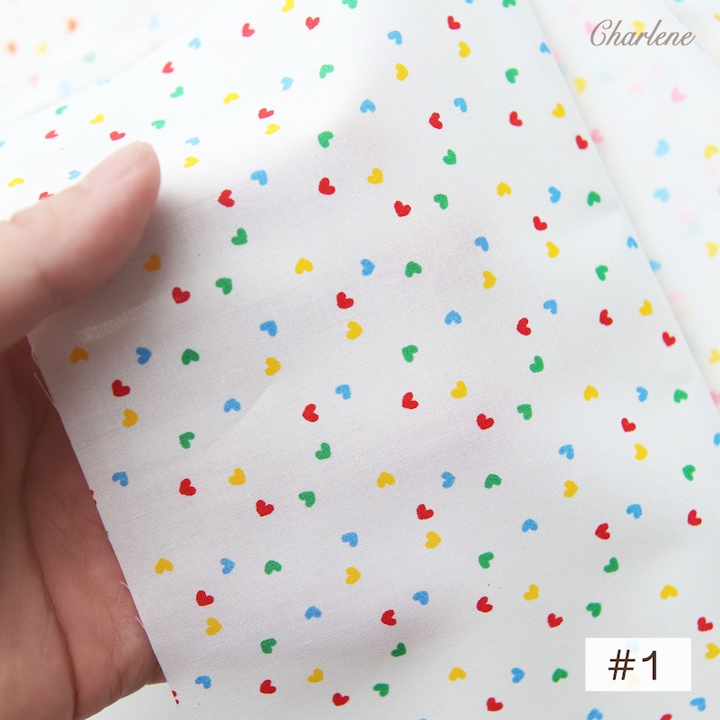 May include: White fabric with a repeating pattern of small, colourful hearts. The hearts are red, yellow, blue, and green. The fabric is folded in half and the text "#1" is visible in the bottom right corner.