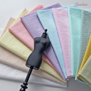 Könnte beinhalten: Eine Sammlung gefalteter Stoffmuster in Pastelltönen wie Gelb, Rosa, Lavendel, Hellblau, Minzgrün und Weiß, die fächerförmig hinter einem schwarzen Schneiderbüsten-Torso angeordnet sind. Das Wort Charlene steht in der oberen rechten Ecke.