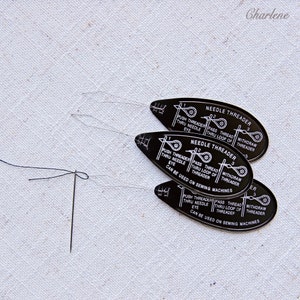 May include: Three black plastic needle threaders with white instructions. The instructions read "Push threader thru needle eye", "Pass thread thru loop of threader", and "Withdraw threader". The threaders are stacked on top of each other. The text "Can be used on sewing machines" is printed on the bottom threader.