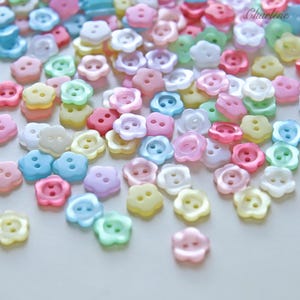 以下が含まれることがあります： ピンク、青、黄色、緑、白など、様々なパステルカラーの5枚花びらの形をした、2つ穴の小さなボタンのコレクション。ボタンは表面に散らばっており、隅にはCharleneという文字が見えます。