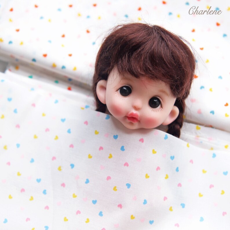 May include: A close-up of a doll's head with brown hair peeking out from a white fabric with a scattered pattern of small pink, blue, and yellow hearts.
