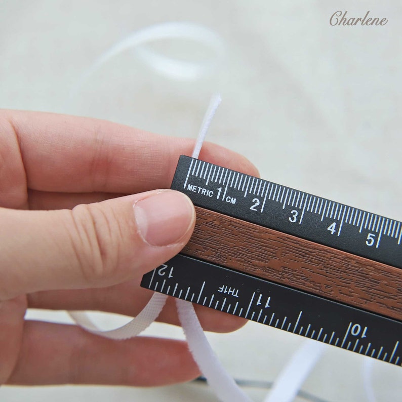 Pu&ograve; includere: Primo piano di un cordoncino elastico bianco misurato con un righello nero. Il righello ha sia misure metriche che imperiali. Il cordoncino elastico &egrave; lungo circa 2,5 cm.