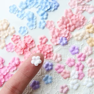 May include: Assortment of small fabric flower petals in various pastel colors, including pink, blue, lavender, and white. The petals are scattered on a white lace background. One white petal is being held by a finger.