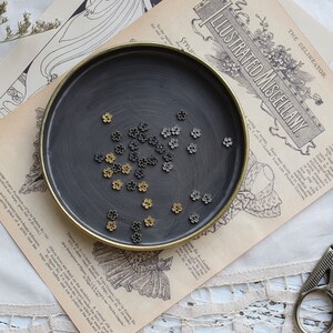 May include: A black metal dish with a gold rim filled with small gold and silver flower-shaped metal charms.