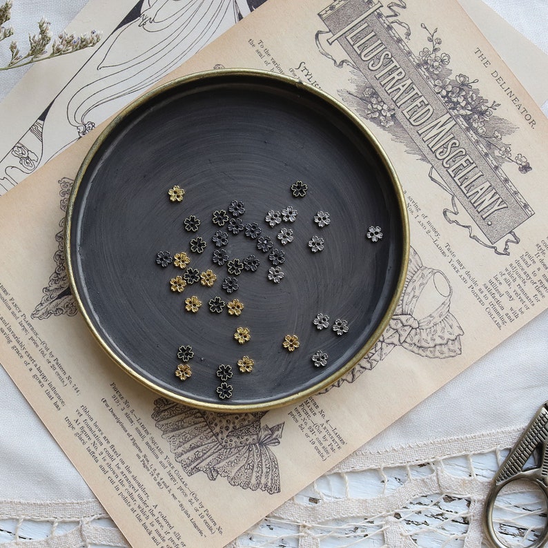 May include: A black metal dish with a gold rim filled with small gold and silver flower-shaped metal charms.