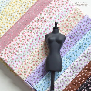 Peut inclure: Un buste de mannequin noir est placé devant plusieurs bandes de tissu pliées, chacune présentant un petit motif floral dans diverses couleurs dont le jaune, le rose, le violet, le bleu et le marron. Le mot Charlene est écrit en haut à droite.