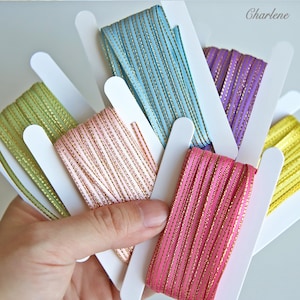 May include: A selection of colourful ribbons, each wound on a white card. The ribbons are in shades of pink, blue, green, purple, and yellow, with gold metallic threads. Ideal for crafts and gift wrapping, the ribbons are presented in a hand.