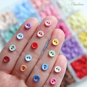 May include: A collection of small, round, two-hole buttons in a variety of pastel and vibrant colours, including pink, blue, yellow, red, orange, and purple, arranged in a grid-like plastic storage container with the name Charlene printed in the corner.