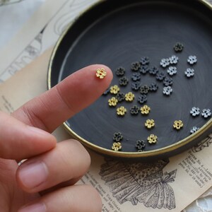 May include: A close-up of a small gold flower-shaped bead. The bead is being held by a finger. There are many other beads in the background, some are gold and some are black.