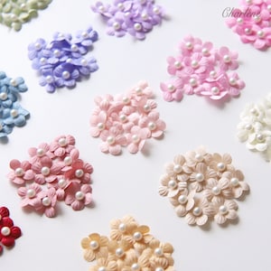 Peut inclure: Des grappes de petites fleurs en papier de différentes couleurs pastel, dont le rose, le lavande et le bleu, sont dispersées sur une surface blanche. Chaque fleur a une perle en son centre. Le nom "Charlene" est visible.