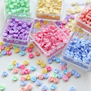 May include: A collection of small, round, two-hole buttons in pastel shades of pink, blue, yellow, purple, and green, organized into clear square containers with loose buttons scattered around, featuring the text Charlene in the corner.