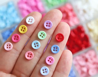 Op de afbeelding: Assortiment van kleurrijke plastic knopen in verschillende kleuren, waaronder rood, blauw, geel, groen, roze en wit. De knopen zijn gerangschikt in een hand, met gespreide vingers.