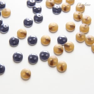 Peut inclure: Une collection de petits boutons ronds à deux trous dans des tons gris foncé et beige translucide, dispersés dans le cadre. Les boutons présentent une finition lisse et polie et sont disposés en motif lâche. Le texte Charlene est visible.