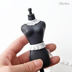 May include: A black mannequin with a white ruffled collar and a white ruffled belt. The mannequin is standing on a white surface.