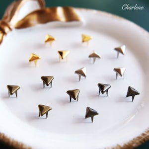 May include: A collection of triangular metal studs in gold, silver, and bronze colors. The studs are arranged on a white plate.