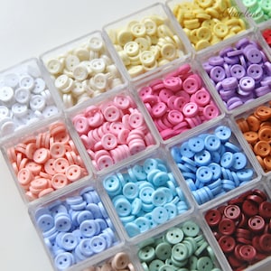 May include: A collection of small, round, two-hole plastic buttons organized by color in a clear, multi-compartment storage container. The buttons are arranged in square sections featuring white, cream, yellow, purple, pink, orange, blue, and red.