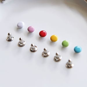 May include: Assortment of colorful round buttons with silver metal bases. The buttons are in a rainbow color order, starting with white, pink, red, yellow, green, and blue.