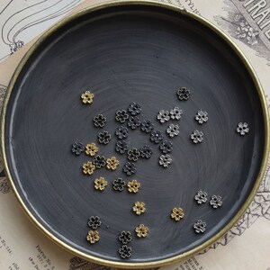 May include: A collection of small metal flower charms in gold, silver, and black colors. The charms are scattered on a dark gray surface.